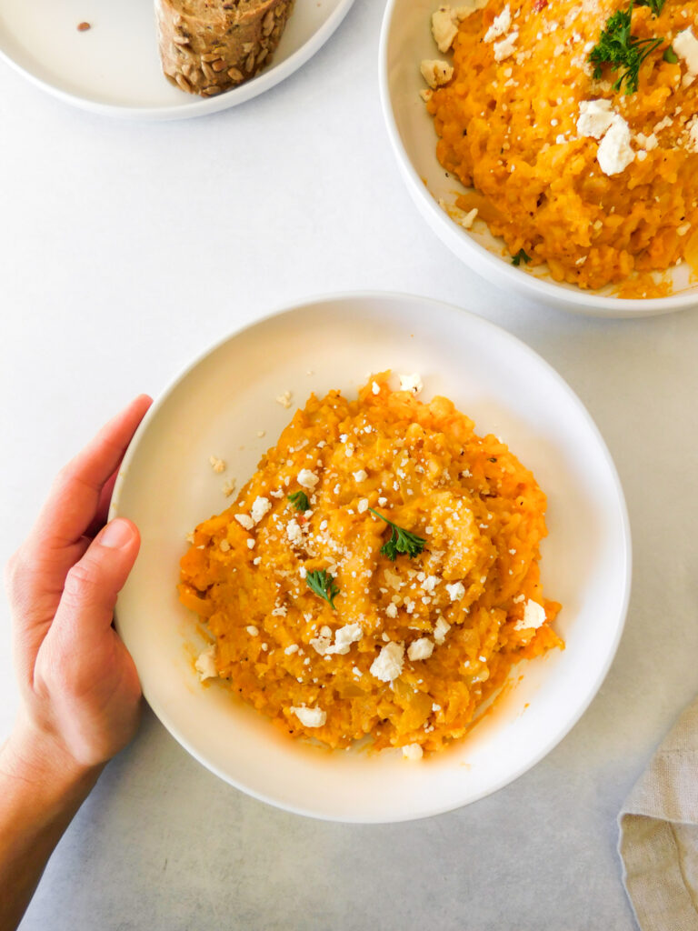 Kaileigh, a white woman, has her hand gently placed around a bowl of roasted butternut squash risotto. Another bowl of risotto sits off to the side and some bread peaks into the frame.
