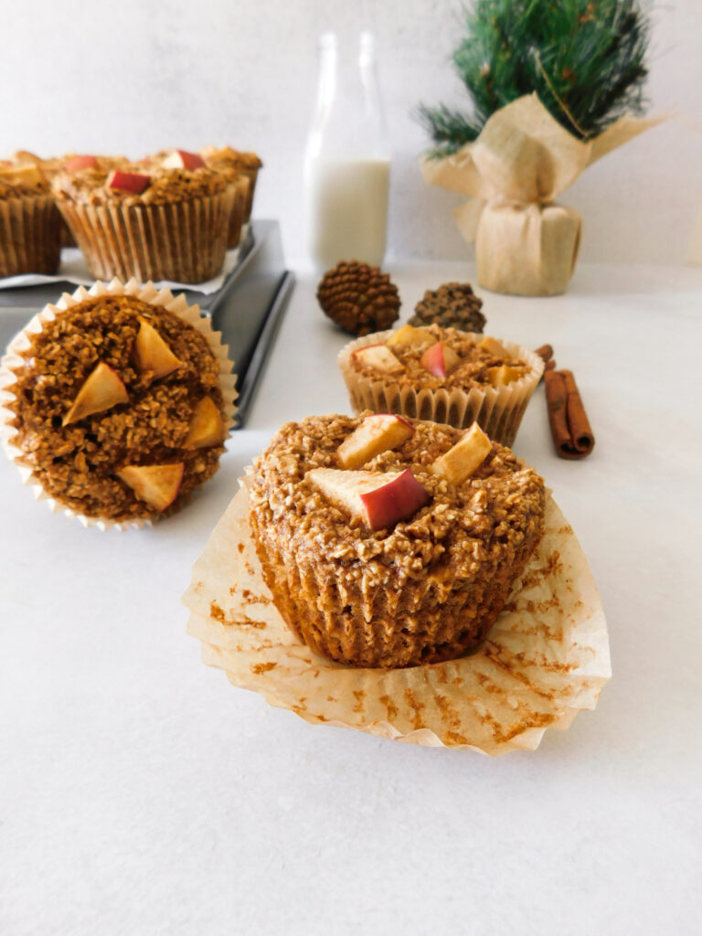 An apple muffin with it's liner opened. More muffins sit behind it as well as a bottle of milk, a small Christmas tree, pinecones, and a cinnamon stick.