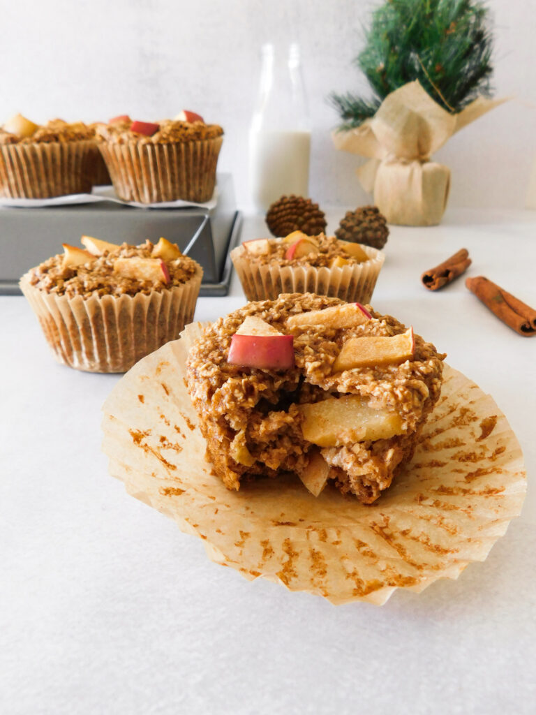 A gluten-free apple protein muffin with a bite out of it. More muffins are sitting behind it as well as a bottle of milk, a small Christmas tree, and cinnamon sticks.