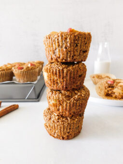 A stack of four Gluten-Free Apple Protein Muffins with several muffins behind it. A bottle of milk is in the background.