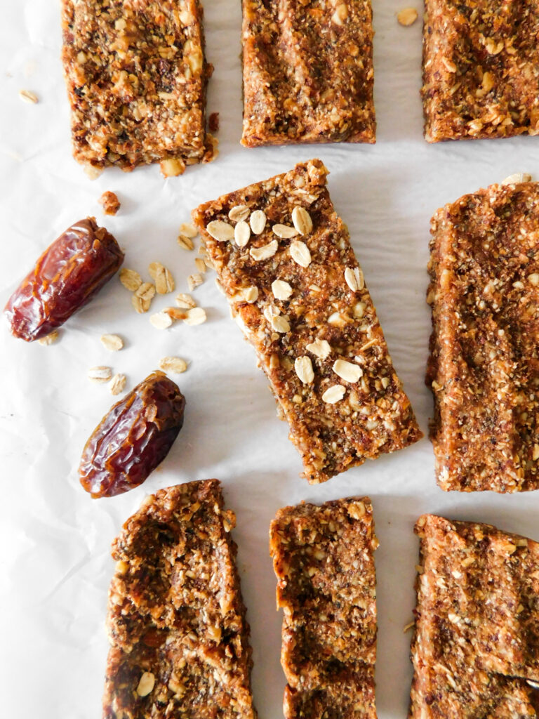 Date bars are in rows with one slightly turned sideways and with oats sprinkled on top of it. Some dates, and oats are scattered next to the energy bars.