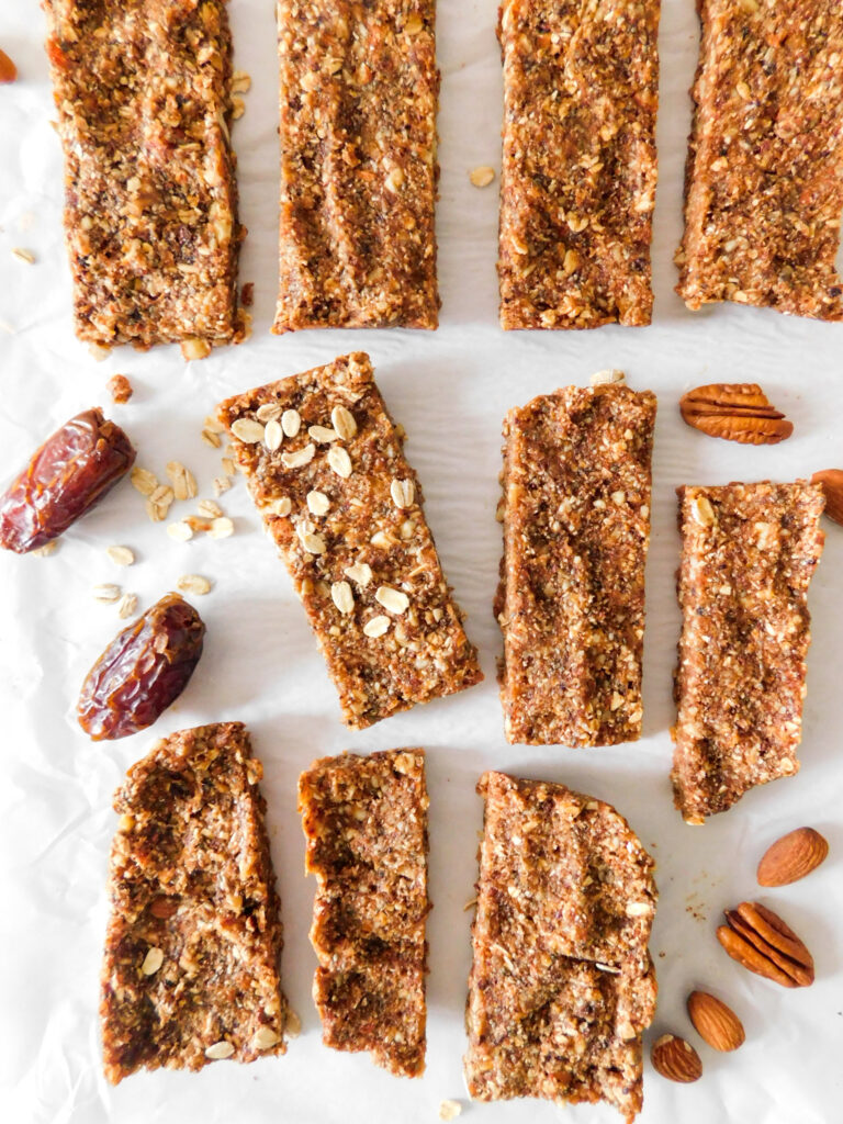 Date bars are in rows with one slightly turned sideways and with oats sprinkled on top of it. Some dates, pecans, and almonds are scattered between the energy bars.