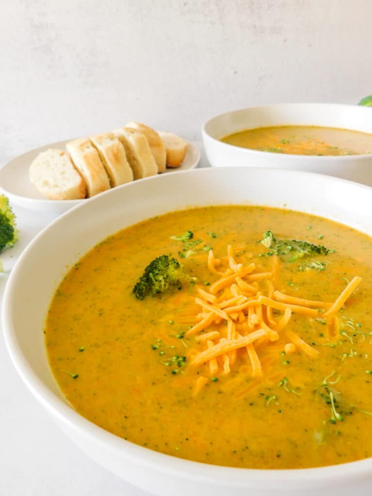 A side view of two bowls of broccoli cheddar soup with some bread slices and broccoli in the background.