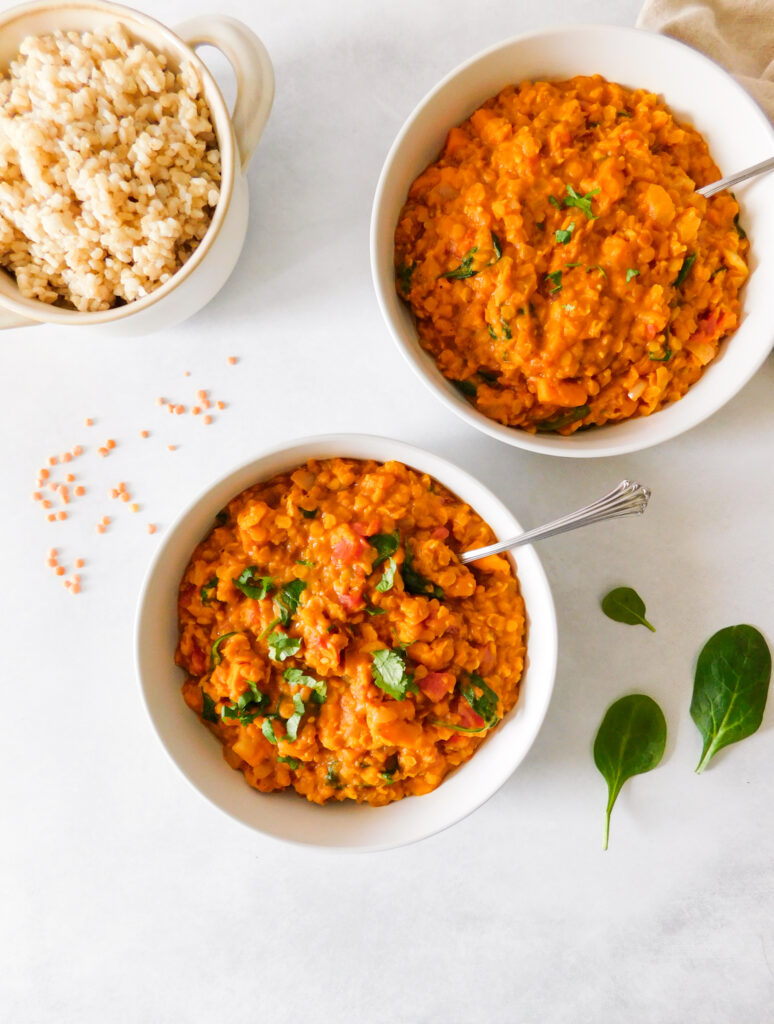 Two bowls of Red Lentil Curry With Sweet Potato and Spinach. A bowl of rice sits to the side. Red lentils are sprinkled on the table and a few leaves of spinach.