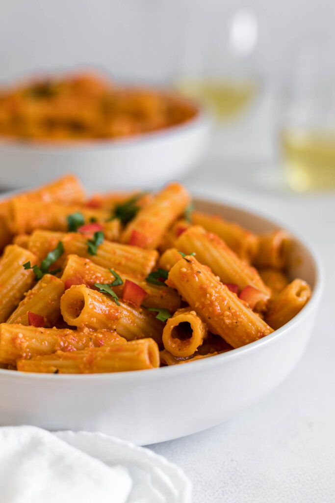 A side view of a bowl of roasted red pepper rigatoni with another bowl in the background. Two white wine glasses are in the background. A white dish towel sits next to the main bowl of rigatoni.