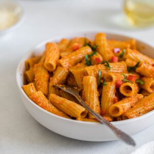 A bowl of Creamy Roasted Red Pepper Rigatoni with another bowl in the background. A glass of white wine sits to the side, as well as a small bowl of parmesan cheese and a white dish towel.