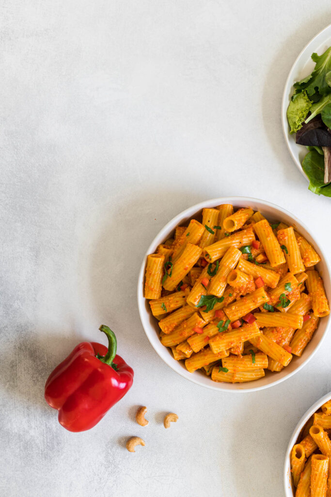 An overhead view of rigatoni pasta in a bowl. A red bell pepper sits next to it along with some cashews.