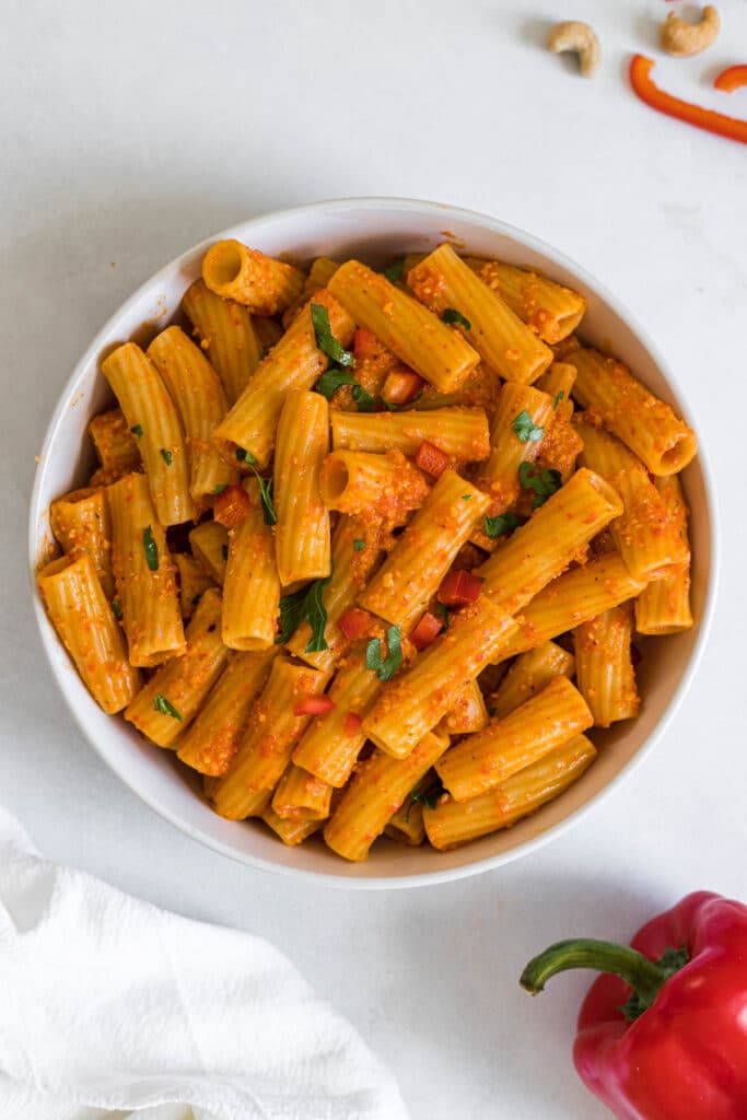 An overhead view of a bowl of rigatoni. A red bell pepper peaks into the frame along with a linen cloth.