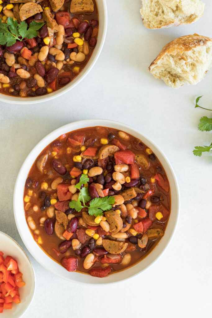 An overhead view of two bowls of vegan chili with two chunks of bread next to them. A small bowl of red bell pepper sits in the bottom left corner and some cilantro is peaking into the frame on the right.
