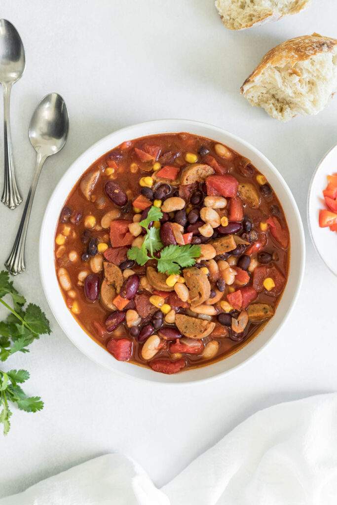 An overhead view of a bowl of vegan chili with two spoons next to it. A chunk of bread and a small bowl of red bell pepper peak into the frame. Some cilantro is peaking into the frame on the left and a linen towel is resting at the bottom of the frame.