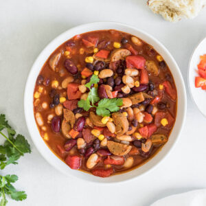 An overhead view of a bowl of Easy 25-Minute Vegan Chili. A chunk of bread and a small bowl of red bell pepper peak into the frame. Some cilantro is peaking into the frame on the left and a linen towel is resting at the bottom of the frame.