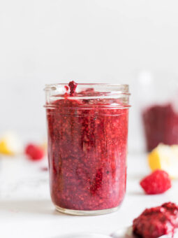 A mason jar with Raspberry Chia Seed Jam in it, with a little spilling out. A spoon with some jam sits in front of the mason jar. Behind the jar is a tablespoon with chia seeds, some raspberries, lemon, and another cup filled with jam.