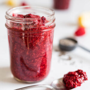 A mason jar with 5-Minute Raspberry Chia Seed Jam in it, with a little spilling out. A spoon with some jam sits in front of the mason jar. Behind the jar is a tablespoon with chia seeds, some raspberries, lemon, and another cup filled with jam.