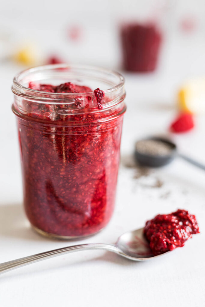 A mason jar with 5-Minute Raspberry Chia Seed Jam in it, with a little spilling out. A spoon with some jam sits in front of the mason jar. Behind the jar is a tablespoon with chia seeds, some raspberries, lemon, and another cup filled with jam.