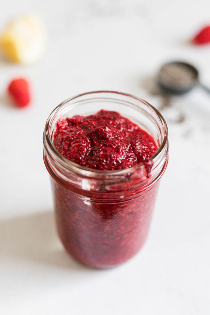 A slightly overhead view of a mason jar with Raspberry Chia Seed Jam. Behind it are some raspberries, a lemon, and a tablespoon with chia seeds.