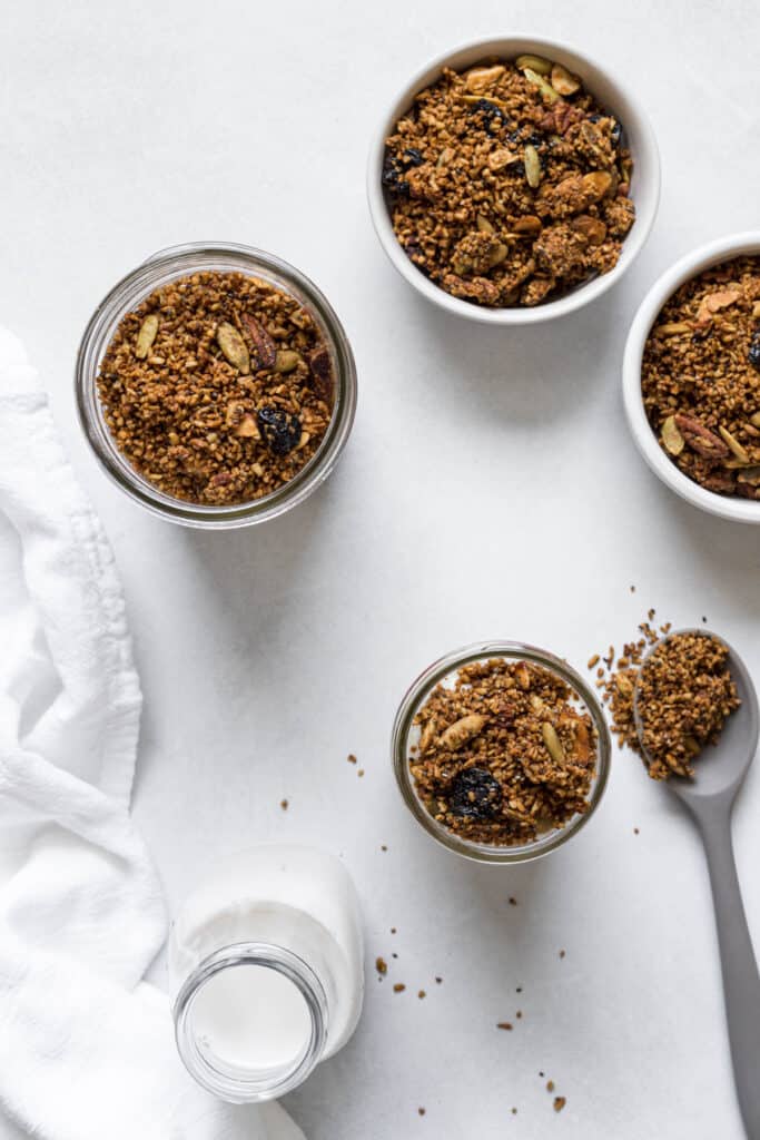 An overhead view of four jars and bowls of steel-cut oat granola. A bottle of milk, linen cloth, and spoonful of granola is on the side.