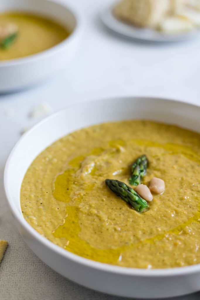 A close up side view of a bowl of asparagus soup with two asparagus tips and white beans in the center of the soup.