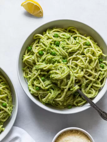 A bowl of Green Pea Pasta with a fork in it. Two lemon wedges and a bowl of parmesan is next to it.