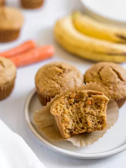 A half eaten Banana Carrot Muffin on a small plate with two other muffins. Bananas and carrots are in the background.