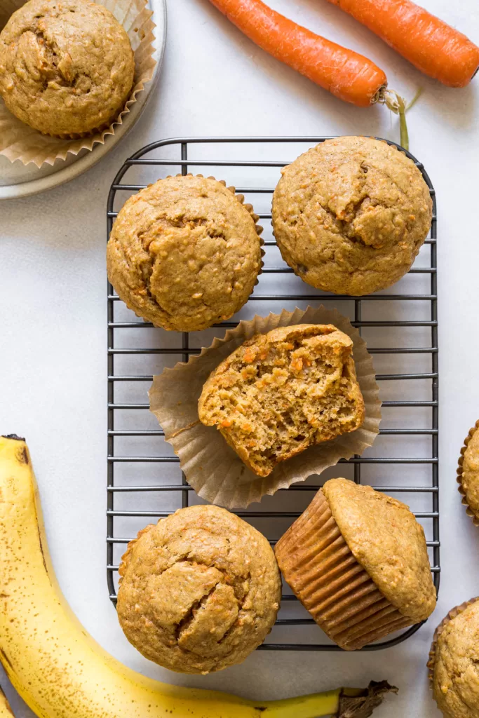 Carrot muffins sit on a wire wrack with carrots and bananas surrounding them.