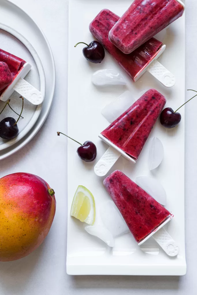 Cherry mango popsicles lay on a long plate with ice cubes, cherries, and a lemon wedge. A mango sits beside it.
