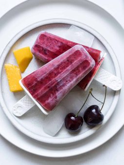 Two Cherry Mango Popsicles sit on a plate with mango cubes and two cherries.