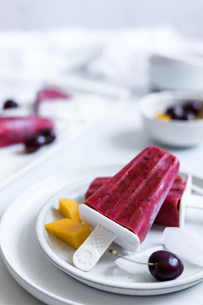 Two popsicles sit stacked on a plate with mango cubes, cherries, and ice cubes next to it. A bowl of cherries is in the background and more popsicles.