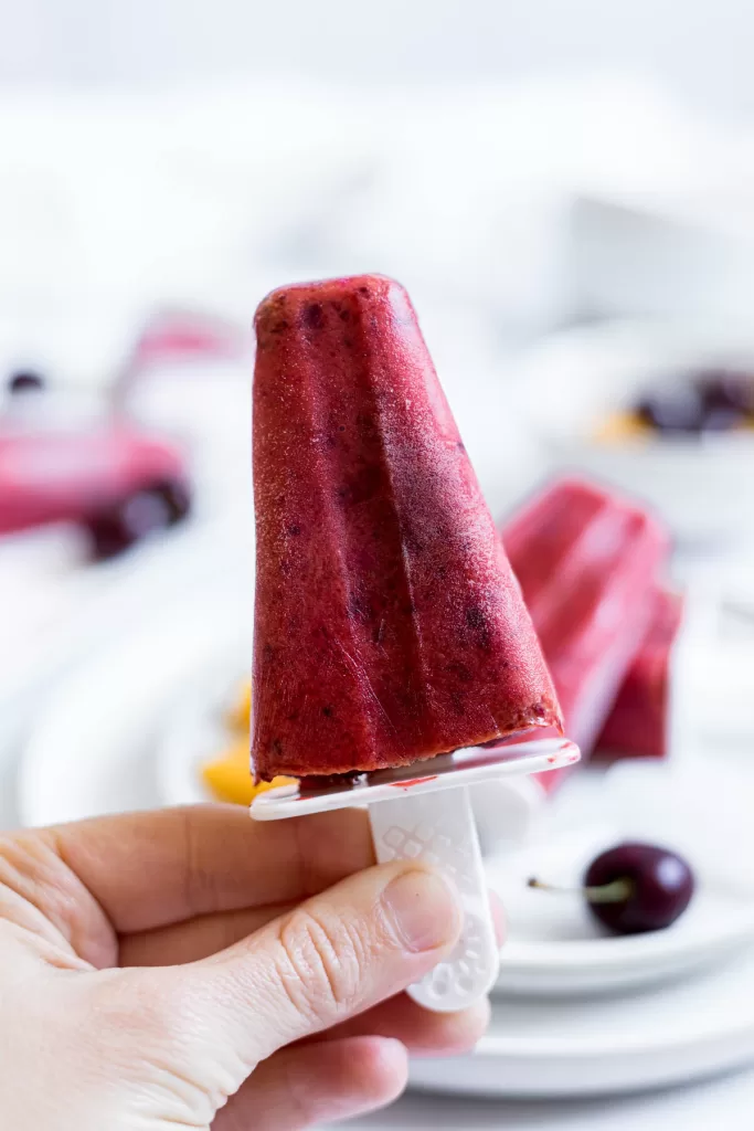 A cherry popsicle is being held up by a white hand. More popsicles are in the background.