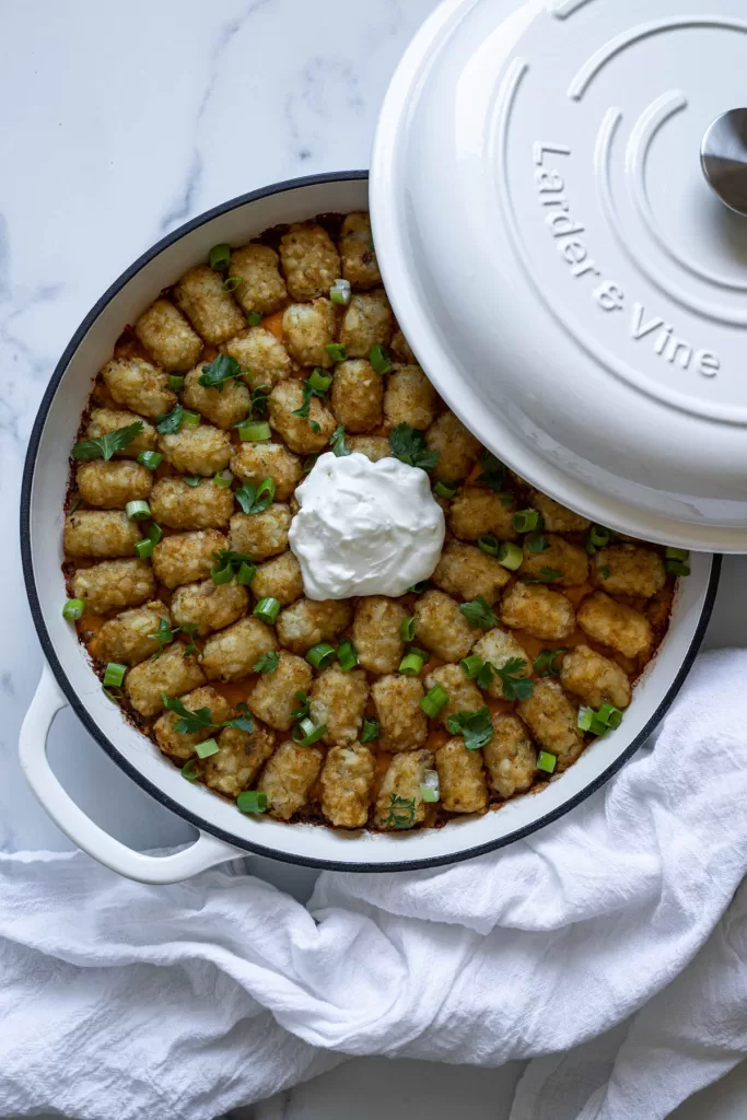 Tater tot casserole in a white braiser with the lid resting on it. A dish cloth sits next to the braiser.
