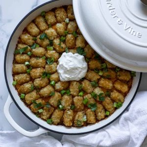 Tater tot casserole in a white braiser with the lid resting on it. A dish cloth sits next to the braiser.