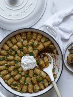 Healthier Tater Tot Casserole sits in a white braiser with a dollop of plain yogurt and green onions on it. A scoop is on a plate and a serving spoon sits in the braiser.