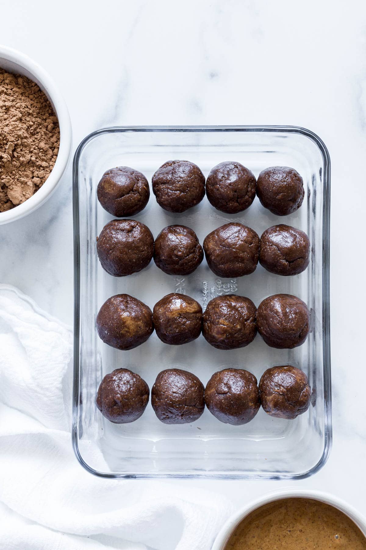 A glass container with rows of Almond Butter Balls and a small bowl of cocoa powder next to it as well as a white dish towel.