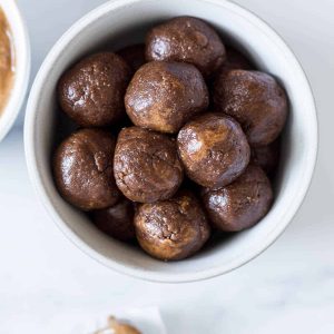 A bowl of Chocolate Almond Balls with a spoon of peanut butter next to it and a bowl of cocoa powder.