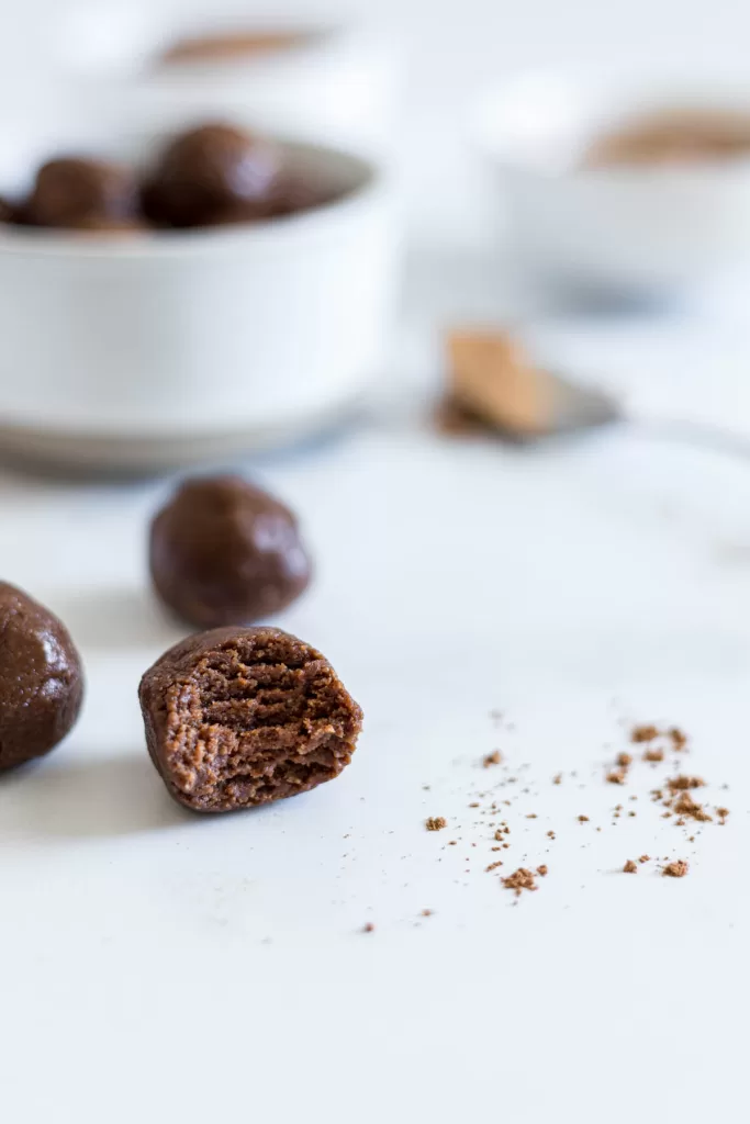 A close up of an Almond Butter Ball with a bite taken out of it and a bowl of chocolate balls behind it.