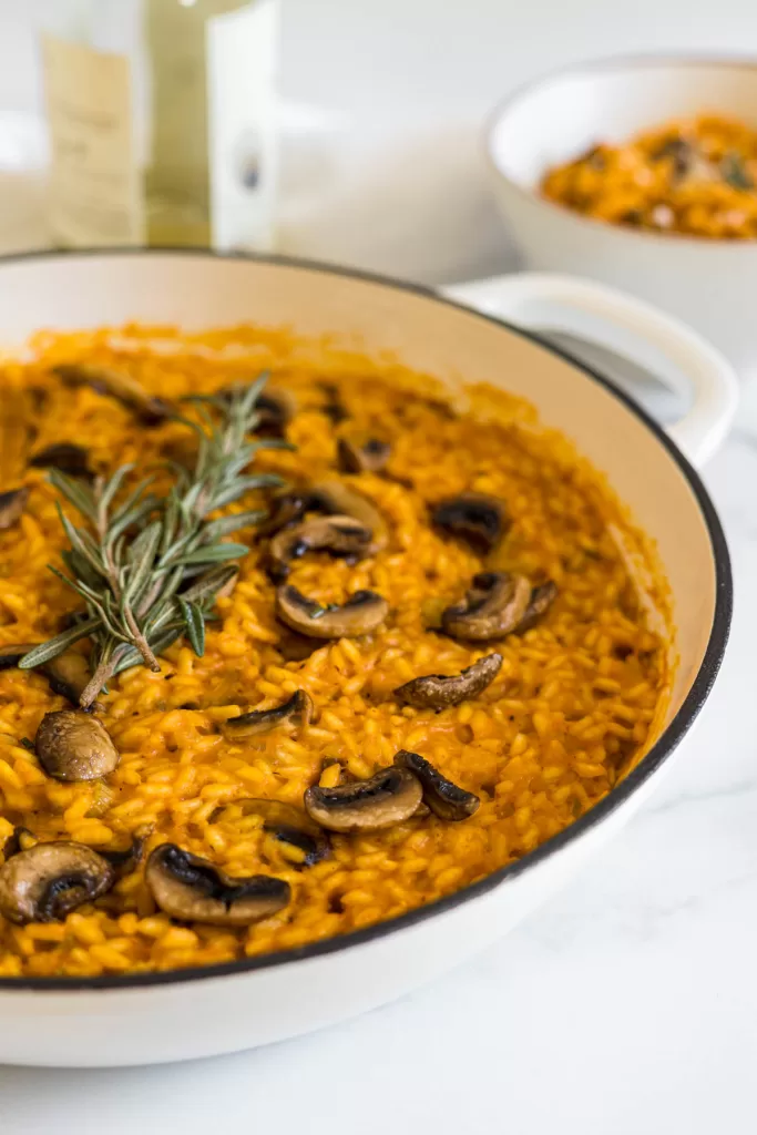 Side view of pumpkin risotto in a braiser with a bottle of wine and a bowl of risotto behind it.
