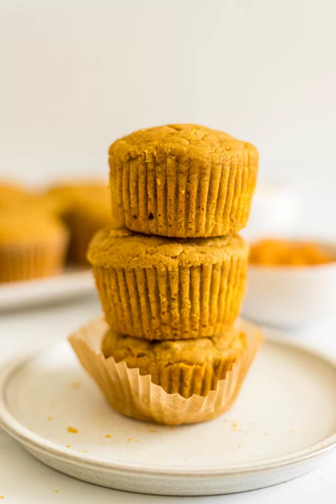 A stack of three pumpkin banana muffins on a plate, the bottom one is in a parchment paper liner. 