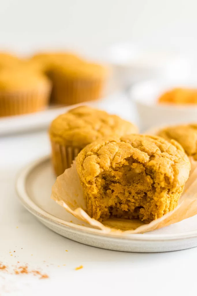 A close up image of a pumpkin muffin with a bite out of it. It's on a plate and sitting in a muffin liner, with more muffins behind it.