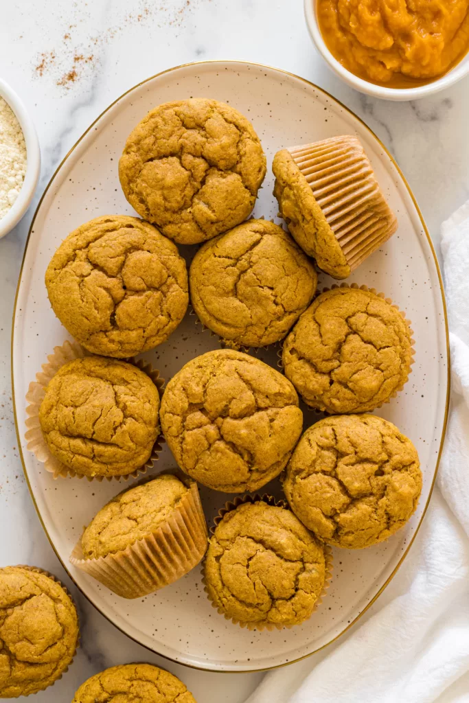 Overhead view of a plate of muffins, two are turned on their sides. Two small bowls of gluten-free flour and pumpkin puree sit next to the plate.