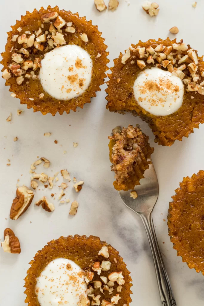 Overhead view of mini pumpkin pies, one with a bite out of it and it's contents are on a fork. Chopped pecans are scattered around.