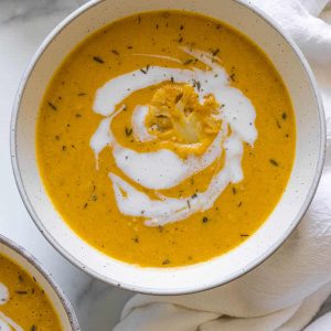 Overhead view of a bowl of pumpkin and cauliflower soup with coconut milk swirled in the soup and a piece of cauliflower set in the middle.
