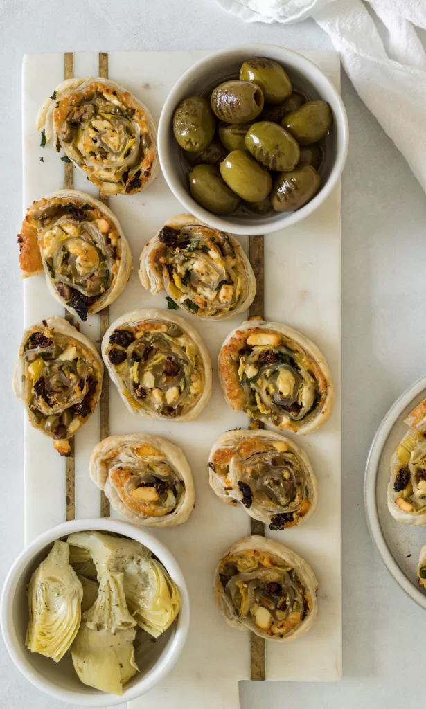 An appetizer marble board with puffed pastry pinwheels and a bowl of artichoke and green olives on it.