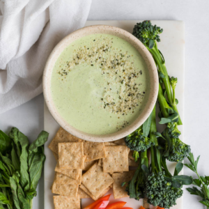 A bowl of green goddess dip with crackers and veggies next to it.
